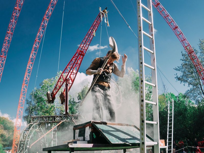 A young man stands on a metal table with a shovel in his hand. Around him are ladders and cranes and some artificial fog.