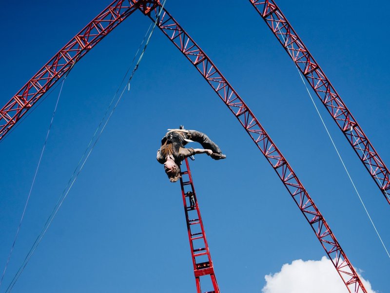 A young man levitating upside down, attached to a crane. There are other crane constructions above him in the sky.