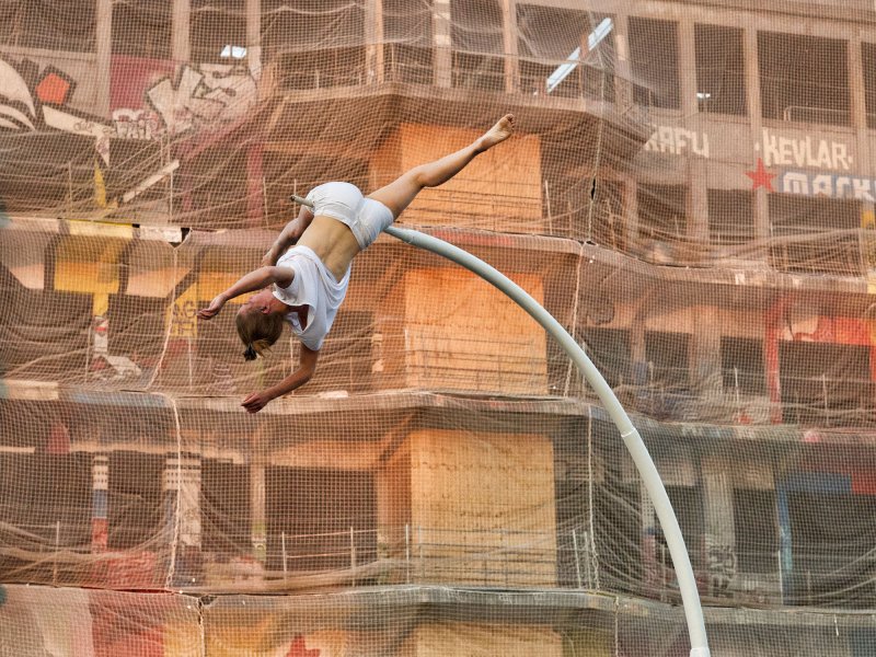 A young woman hanging acrobatically upside down from a curved pole. In the background is a multi-storey building covered with nets.
