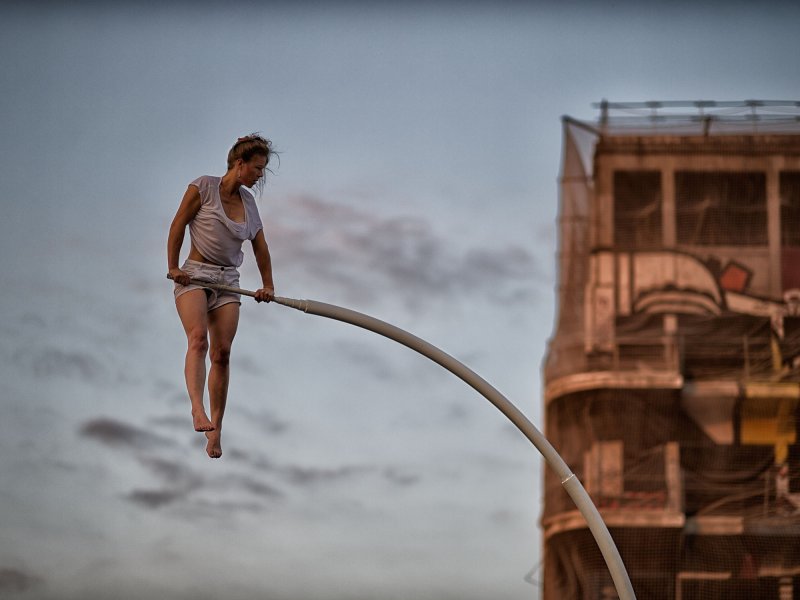 A young woman pushing herself up with her hands from a curved bar. In the background is a multi-storey building covered with nets.