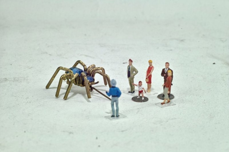 Different sized and colourfully dressed miniature people look at a golden coloured giant spider.