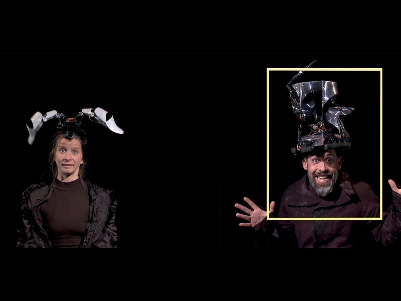 Ausschnitt von zwei Menschen vor einem schwarzen Hintergrund. Links steht eine Frau mit elektrischen Hasenohren auf dem Kopf. Rechts von ihr steht ein Mann, der einen zylinderartigen Hut aus Metall und Elektronik trägt und lächelt. Er wird von einem gelben Rechteck umrandet.