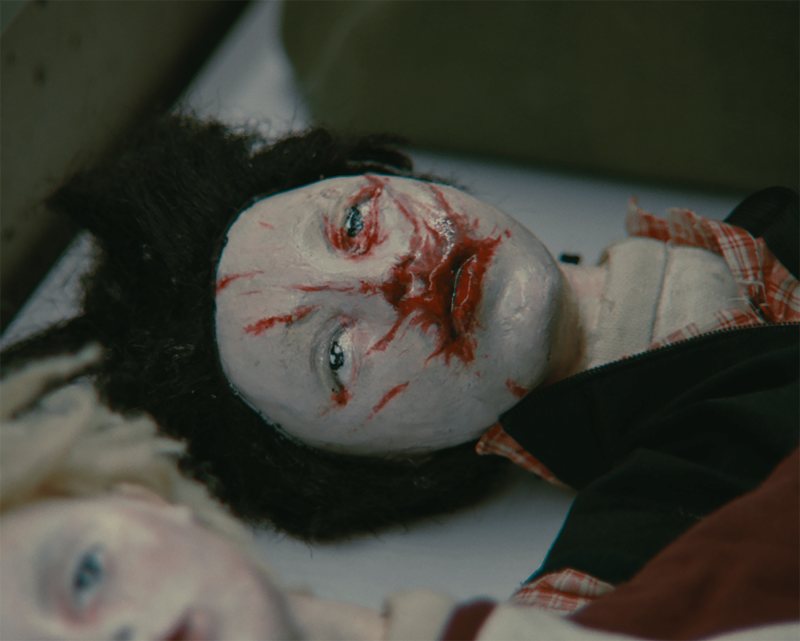 The head of a doll with black hair and twisted eyes. The face is sprayed with red paint. It looks like the doll is bleeding out of its mouth.