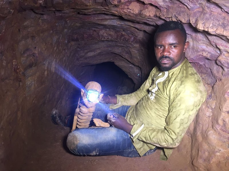 Ein Mann sitzt auf dem sandigen Boden eines engen Tunnels. In der Hand hält er eine Holzpuppe, die eine Stirnlampe trägt.