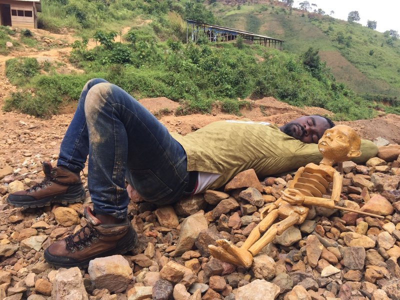 A man is sleeping on a rocky ground. Next to him lies a wooden doll. In the background is a green hill with a barrack on its slope.