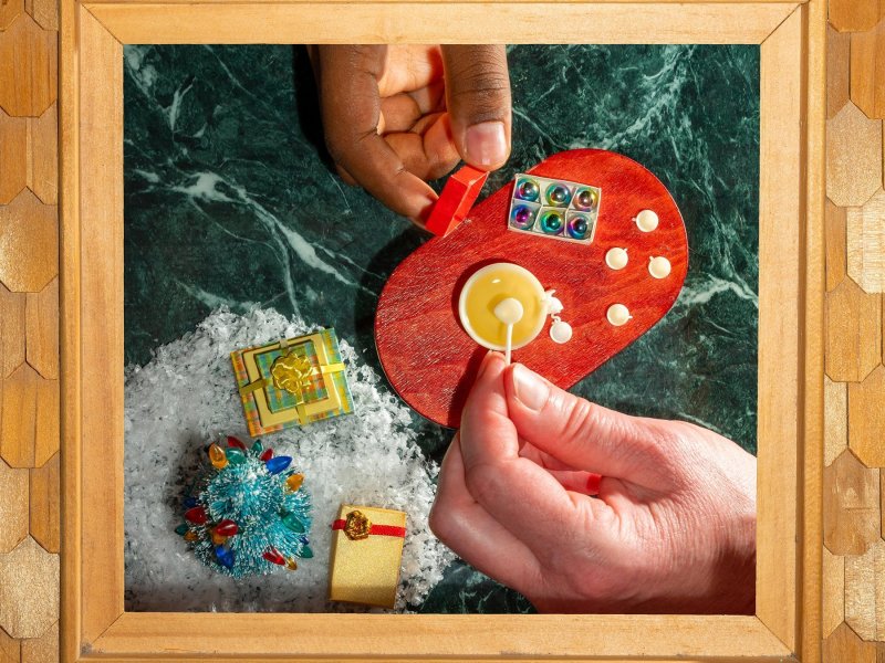 A framed picture of a miniature Christmas from above, played with by two hands. On the left a decorated tree and presents lying on artificial snow, on the right an oval wooden table on which are five small cups, a punch bowl and a box of Christmas balls.