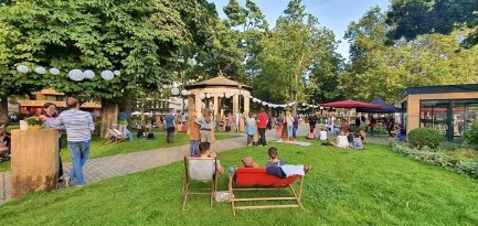 Menschen sitzen und stehen in einem Park mit Bäumen, Lampions und einem Pavillon bei Tageslicht