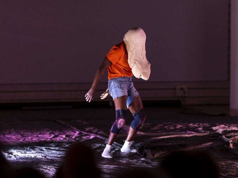 Eine Person in einem orangenen T-Shirt, einer Jeansshorts und Knieschonern gekleidet tanzt auf einer mit schwarzer Plastikplane verdeckten Bühne. Das Gesicht der Person ist von einer Teigmasse bedeckt.