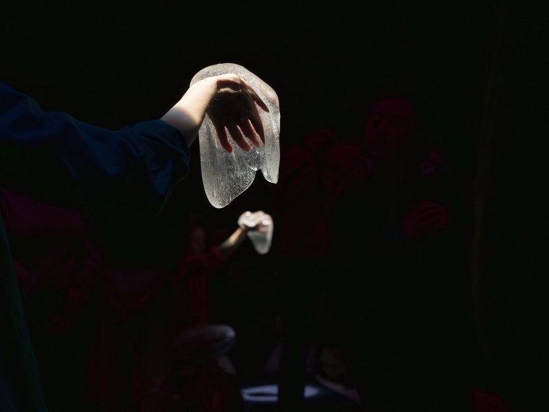 Im Vordergrund ist eine Hand zu sehen, die eine eine transparente, formbare Masse in einer dunklen Umgebung hält. Die Hand und die Masse werden von hellem Licht angestrahlt. Im Hintergrund ist undeutlicher eine weitere hell angestrahlte Hand mit Masse zu sehen.