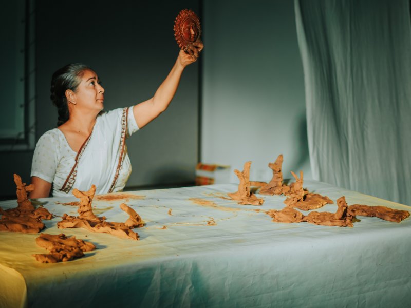 A woman in traditional dress holds up a moulded clay object while she works with other clay figures at the table. The scene is softly lit.