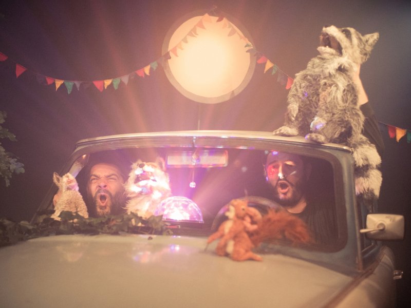 Zwei Männer im Auto, das von Pflanzen umgeben ist, mit Tierpuppen und einer Kugel, die buntes Licht ausstrahlt. Über dem Auto hängen bunte Girlanden und ein Mond ist zu sehen.