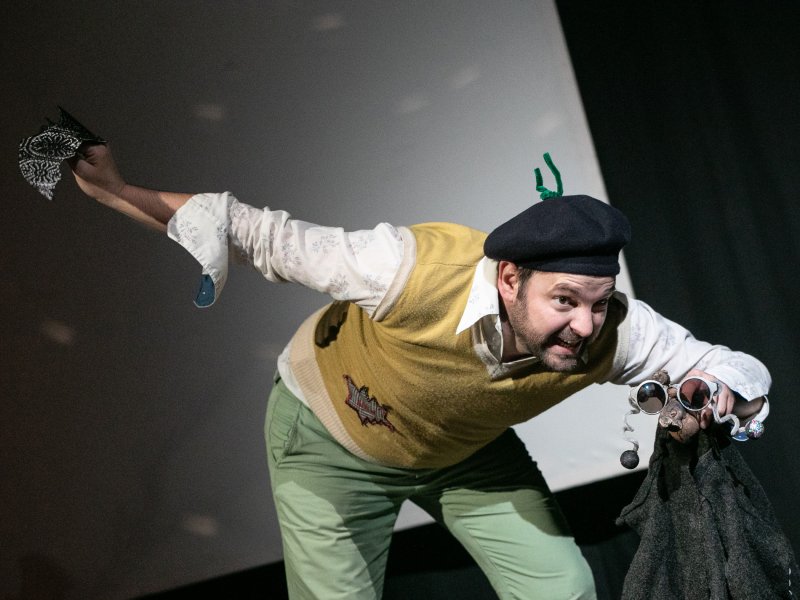 Schauspieler in einem gelben Pullover und grüner Hose, der dynamisch auf einer Bühne posiert. Er trägt eine schwarze Mütze, hält eine schwarze Jacke und eine Sonnebrille. Der Gesichtsausdruck ist lebhaft und ausdrucksvoll.