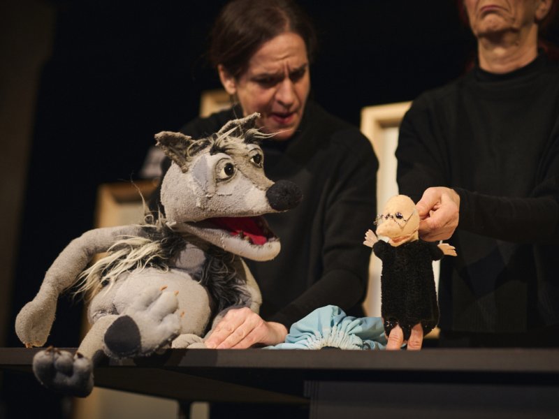 Eine Person hält eine große wolfähnliche Puppe und eine andere Person zeigt eine kleine schwarze menschenähnliche Puppe mit Brille, großer Nase und mit beweglichen Armen. Der Hintergrund ist schwarz.