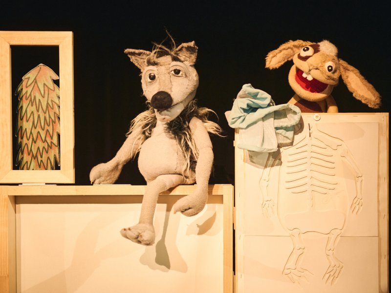 Puppet show with two figures: a grey wolf and a brown hare. The wolf sits on a box, the hare stands behind a wooden panel on which a small skeleton has been carved.