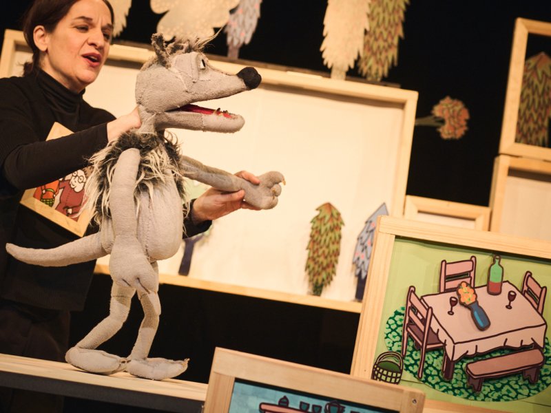 Puppeteer performs a gray wolf puppet on stage. Painted backdrops with furniture and plants are visible in the background.