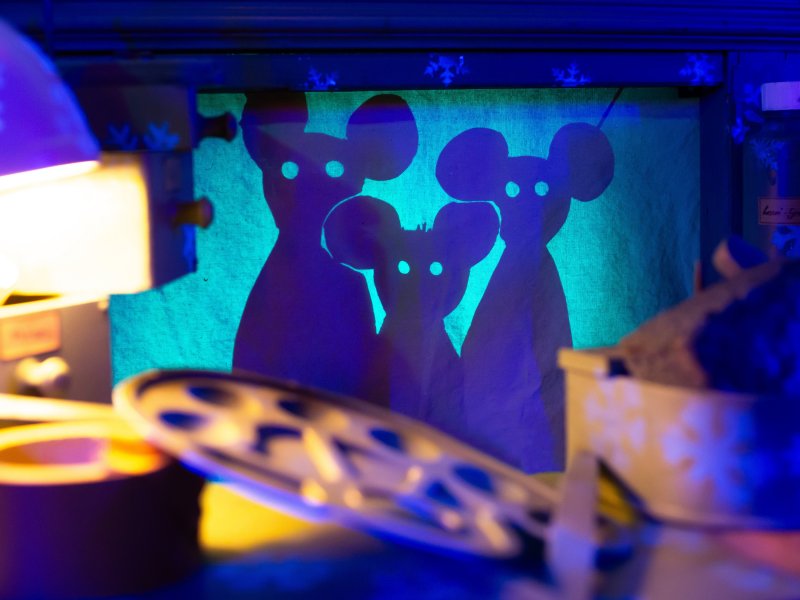 Schatten von drei Mäusen, die auf einer transparenten Papierwand zu sehen sind. Im Vordergrund liegen verschiedene Materialien und eine Filmrolle auf einem Tisch.