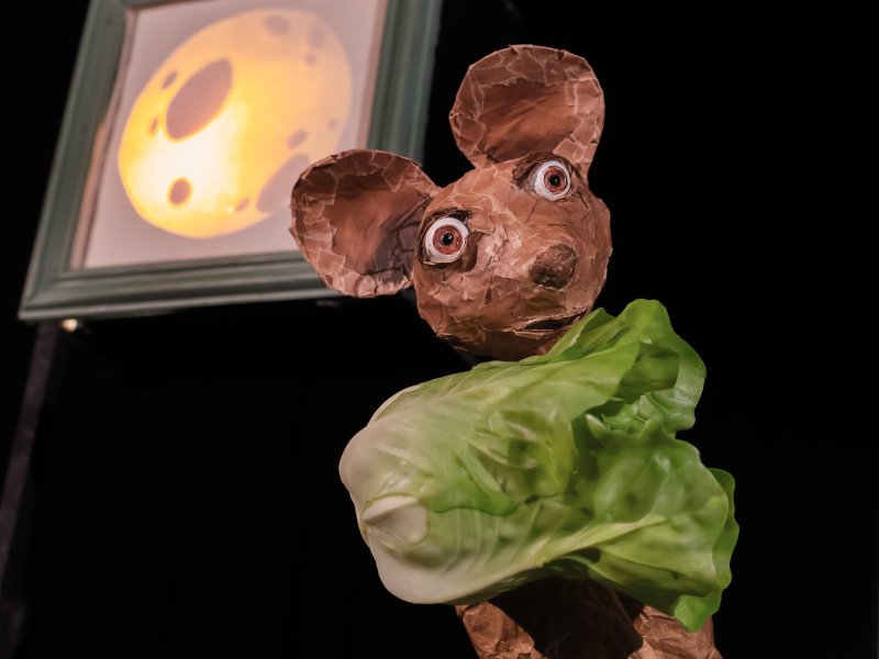 Papier-mâché mouse with green leaves in the foreground in front of a picture of a moon in the background.