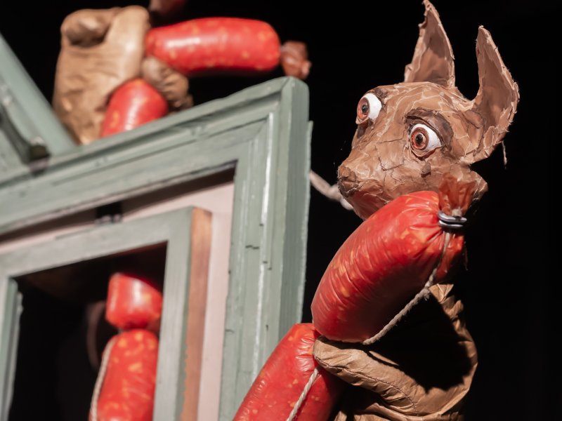 A papier-mâché mouse holding a red sausage chain and peering out from behind a window frame. In the background, another doll, only partially visible, also clutching the sausage chain.