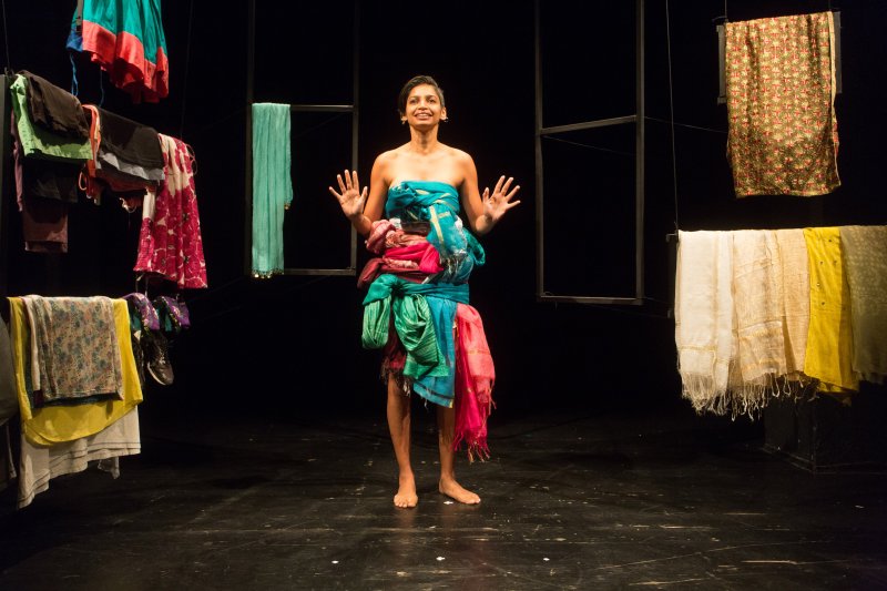 Woman with her hands raised at her sides, dressed in colourful knotted scarves. She stands in the middle of a dark stage, surrounded by poles with more colourful scarves and dresses.