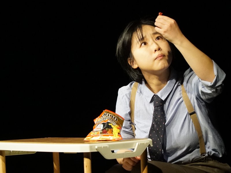 Actress in a shirt and tie sitting at a table, looking thoughtfully at her raised hand in which she is holding a gummy bear. There is a packet of Haribo gummy bears on the table.