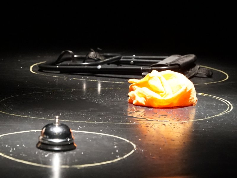 An orange-coloured cloth, a table bell and a folded chair lie on a black floor. All three objects are each surrounded by a brightly coloured circle.