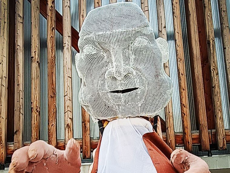 Große Drahtskulptur eines menschlichen Kopfes mit übergroßen Händen, vor einer Holzlattenwand