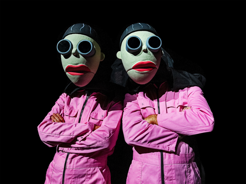 Zwei Personen in pinken Anzügen mit Puppenmasken, mit dicken, roten Lippen und verdunkelten Brillen.