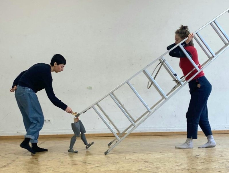 Zwei Personen in einem leeren Raum, eine hält eine Leiter, die andere führt eine kleine Figur mit Leiter, die aussieht, als würde sie die große Leiter tragen.