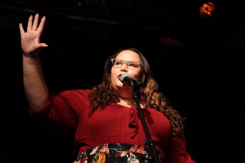 Auf dem Bild ist eine Frau in einem roten Oberteil an einem Mikrofon zu sehen, die einen Arm erhoben hat und den Horizont zeigt.