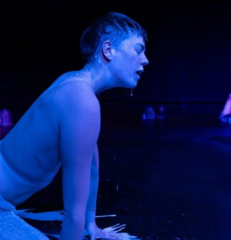 Eine Person mit kurzem Haar kniet auf dem Boden und stützt sich mit den Händen ab. Die Person trägt ein ärmelloses Oberteil und ist von blauem Licht beleuchtet. Vom Kinn der Person tropfen Flüssigkeitstropfen nach unten. Der Hintergrund ist dunkel.