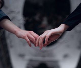 Zwei Hände, die sich leicht berühren, wobei die Finger sich sanft ineinander verschränken. Die Hände sind vor einem unscharfen, dunklen Hintergrund zu sehen. Eine Hand trägt einen dunklen Ärmel, die andere einen helleren Ärmel.