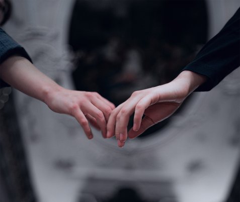 Zwei Hände, die sich leicht berühren, wobei die Finger sich sanft ineinander verschränken. Die Hände sind vor einem unscharfen, dunklen Hintergrund zu sehen. Eine Hand trägt einen dunklen Ärmel, die andere einen helleren Ärmel.