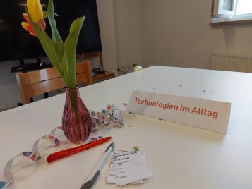 Tisch mit Blumenvase, Zetteln und Stiften. Schild mit Aufschrift Technologien im Alltag