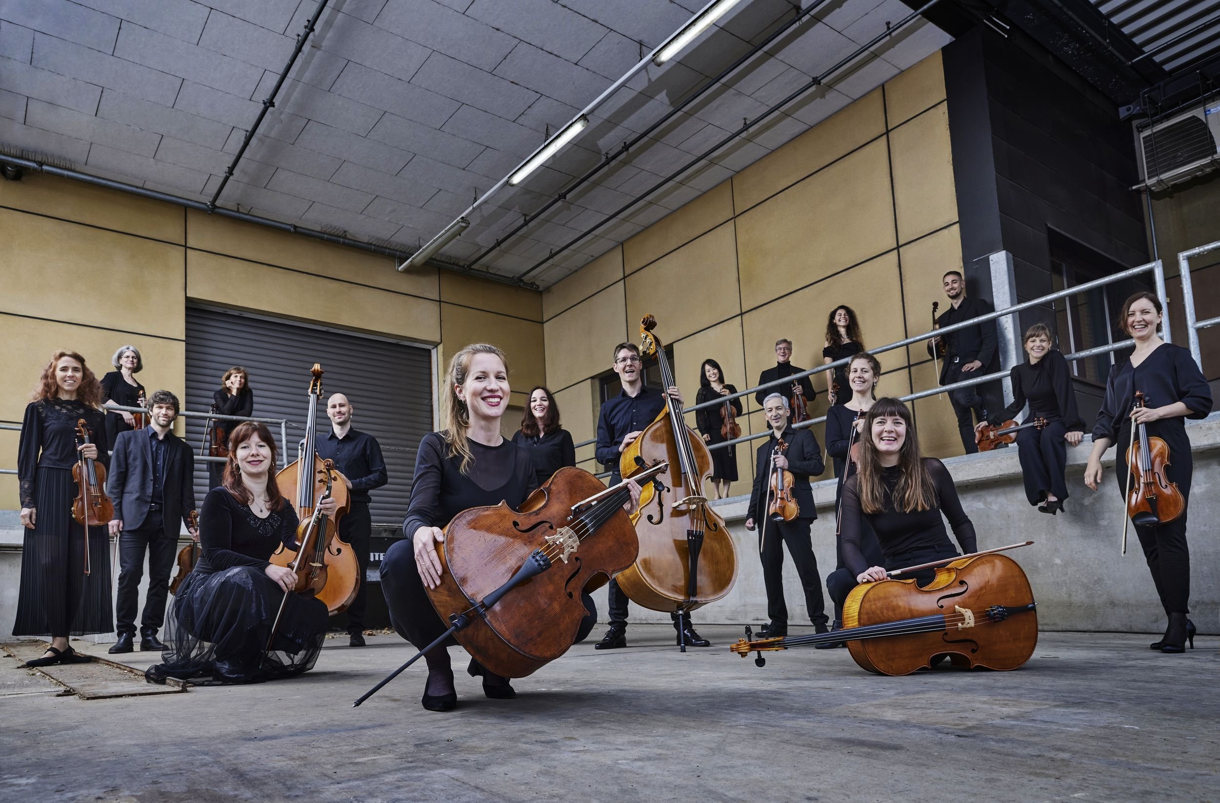 Gruppe von Musikerinnen und Musikern mit Streichinstrumenten wie Cello und Violine in einem modernen, industriellen Innenhof.