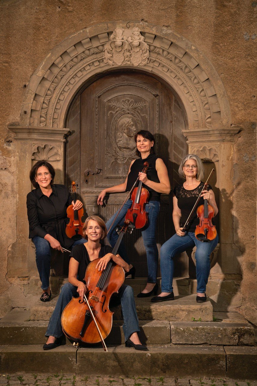 Vier Musikerinnen mit Streichinstrumenten (drei Geigen, ein Cello) sitzen und stehen vor einem verzierten, steinernen Torbogen mit einer Holztafel in der Mitte.