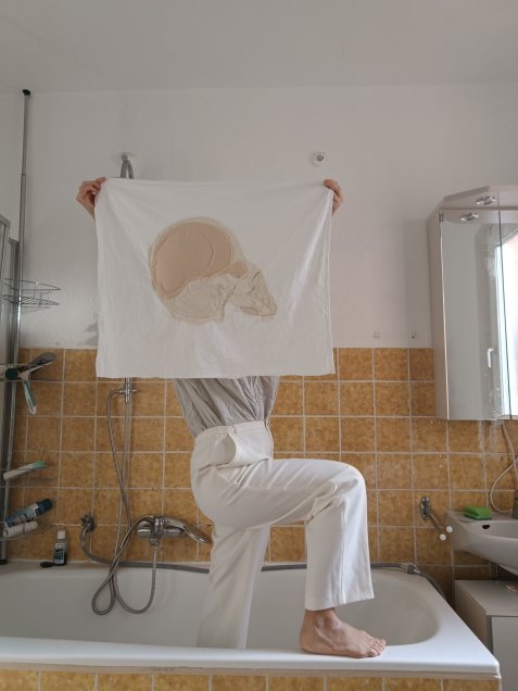 Person hält ein mit einem Schädel in Weiß und Beige besticktes Tuch in die Höhe, sodass das Gesicht der Person verdeckt wird