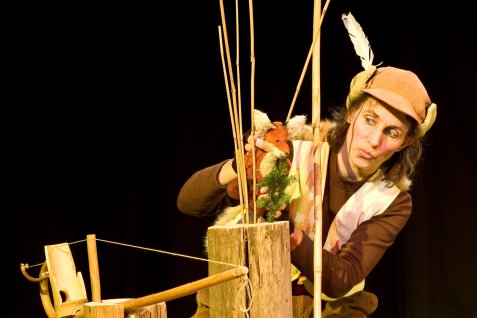Person mit braunem Oberteil und brauner Mütze mit weißer Feder hält eine Fuchs-Handpuppe über eine Holzstruktur mit Stäben.
