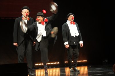 Drei Personen in schwarzer Clowns-Kleidung stehen auf einer Bühne mit dunklem Hintergrund. Die mittlere Person springt und bewegt ihre Arme. Die anderen beiden Personen stehen seitlich und ruhig.