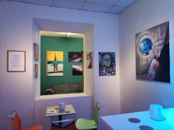 Kleiner Ausstellungsraum mit mehreren Gemälden an den Wänden, darunter ein Bild mit Erde und ein Sofa im angrenzenden Raum.