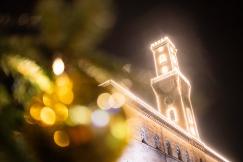 Das festlich beleuchtete Rathaus mit hervorgehobenem Turm, unscharf im Vordergrund Weihnachtsbaumkugeln mit Lichtern.