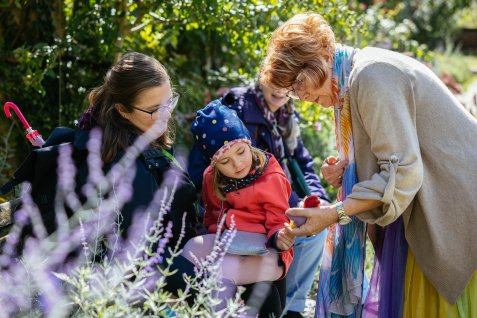 Märchenerzählerin Zorica Otto zeigt einem Kind im Stadtpark etwas in der Hand, umgeben von weiteren Personen und Pflanzen.