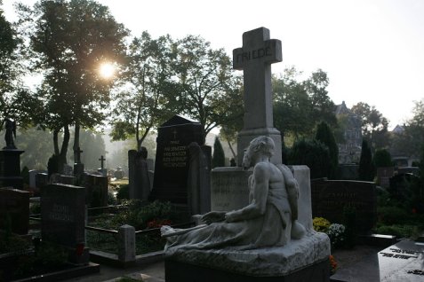 Grabstein mit einer liegenden Skulptur vor einem Kreuz mit der Inschrift 'FRIEDE' auf einem städtischen Friedhof bei Sonnenlicht und Bäumen im Hintergrund.