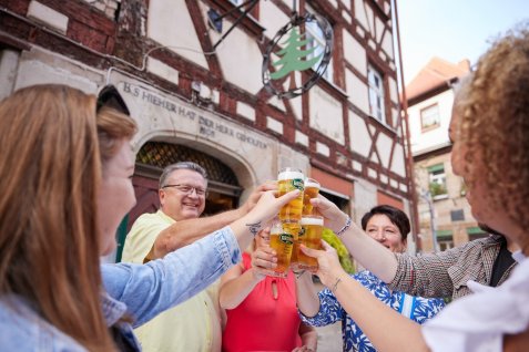 Mehrere Personen stoßen mit Biergläsern vor einem Fachwerkhaus an, das eine Gaststätte ist.