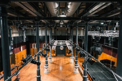 Große Halle des Kulturforums mit Holzboden, schwarzen Säulen und einer zentralen Beleuchtungs- und Lautsprecheranlage an der Decke.