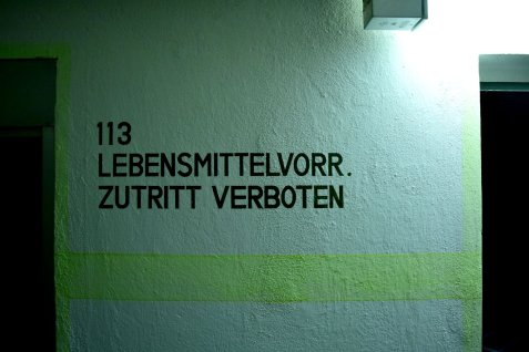 Schwarze Wandbeschriftung mit der Nummer '113' und dem Text 'LEBENSMITTELVORR. ZUTRITT VERBOTEN' auf einer hellen Wand im Kronacher Bunker.