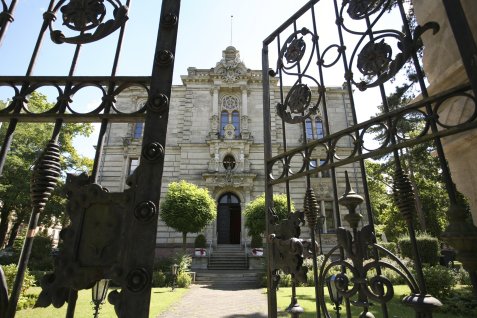 Blick durch ein geöffnetes schmiedeeisernes Tor auf den Eingang des Logenhauses mit Treppe und verzierten Fenstern, umgeben von Bäumen und Garten.
