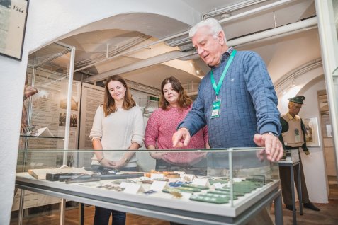 Drei Personen stehen vor einer Vitrine mit verschiedenen Exponaten im Kriminalmuseum, eine Person zeigt auf die Ausstellungsstücke.