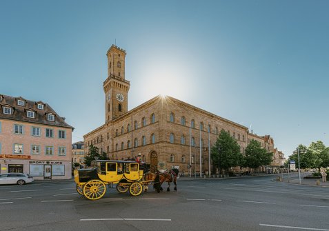 Gelbe Postkutsche mit zwei Pferden vor dem historischen Rathaus mit hohem Turm in der Sonne.