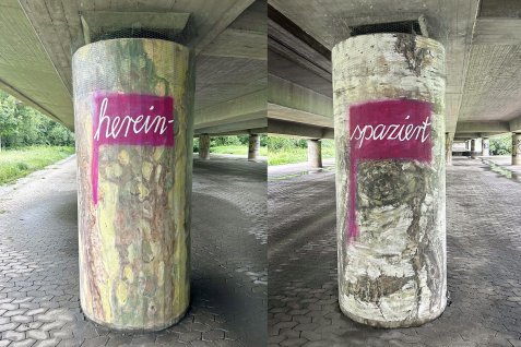 Zwei Brückenpfeiler der Ludwigsbrücke, bemalt von Sabine Deifel als Baumstämme mit den weißen Schriftzügen 'herein-' und 'spaziert' auf rotem Hintergrund.