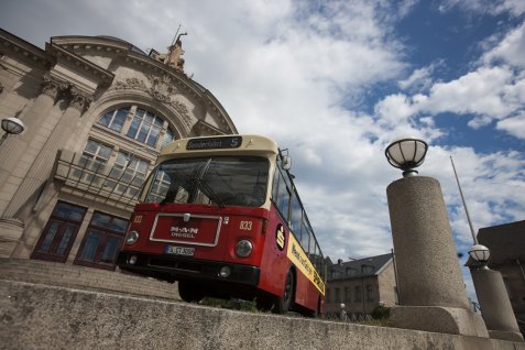 Alter roter Bus mit der Aufschrift 'Sonderfahrt' steht vor einem historischen Gebäude mit großen Fenstern und Säulen. Im Vordergrund sind zwei runde Straßenlaternen auf Betonpfeilern zu sehen.